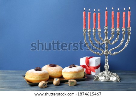 Silver menorah, dreidels with He, Pe, Nun, Gimel letters, gift box and sufganiyot on blue wooden table, space for text. Hanukkah symbols