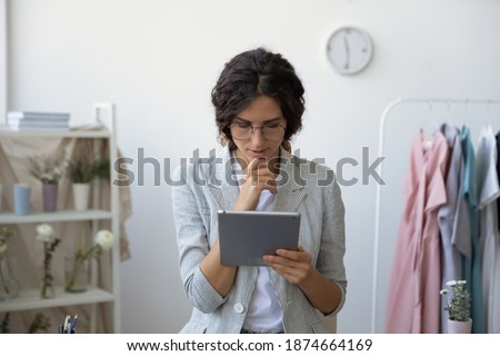 Concentrated millennial businesswoman designer standing at studio atelier reading email from digital tablet screen. Self employed private enterpreneur pondering on answer to customer client message Royalty-Free Stock Photo #1874664169