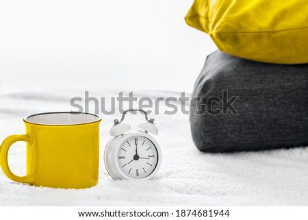 Illuminating Yellow Cup or mug with tea or coffee, Ultimate Gray alarm clock and pillows with colors of the year 2021 on bed in bedroom. Trendy good morning and home decor