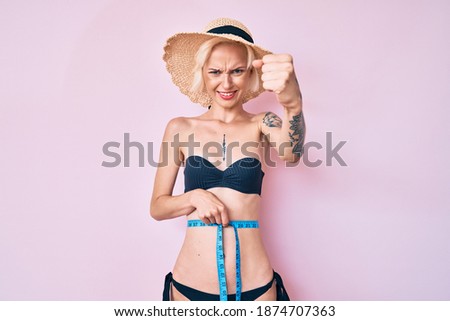 Young blonde woman with tattoo wearing bikini using tape measure annoyed and frustrated shouting with anger, yelling crazy with anger and hand raised 