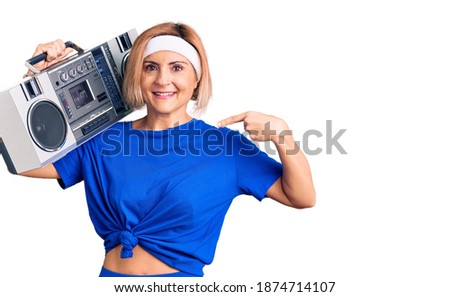 Young blonde woman wearing sportswear holding boombox, listening to music pointing finger to one self smiling happy and proud 