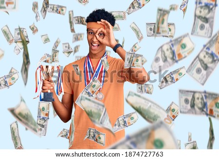 Young african american man holding champion trophy wearing medals smiling happy doing ok sign with hand on eye looking through fingers