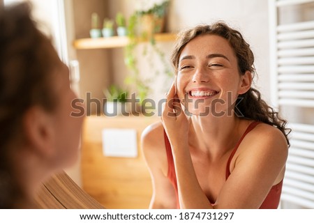 Happy smiling beautiful girl cleaning skin face with cotton pad. Beauty natural woman looking in mirror while cleansing skin face and using cosmetic products for properly deep clean. Royalty-Free Stock Photo #1874731297