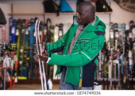 Positive African American man choosing ski in store of sports equipment.
