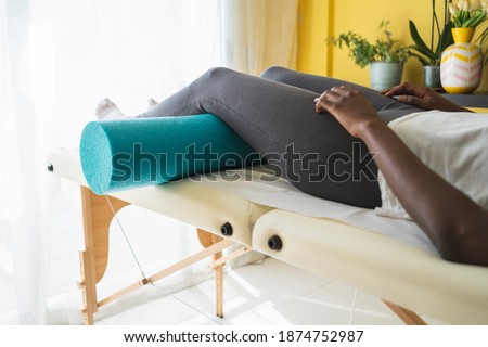 Legs of an african woman in tights using a foam cylinder to do physical therapy exercises while lying on a stretcher in a clinic Royalty-Free Stock Photo #1874752987