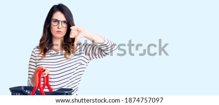 Beautiful young brunette woman holding supermarket shopping basket with angry face, negative sign showing dislike with thumbs down, rejection concept 