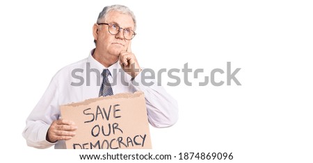 Senior grey-haired man wearing business clothes holding save our democracy protest banner serious face thinking about question with hand on chin, thoughtful about confusing idea 