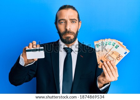 Attractive business man with long hair and beard holding credit card and euros relaxed with serious expression on face. simple and natural looking at the camera. 