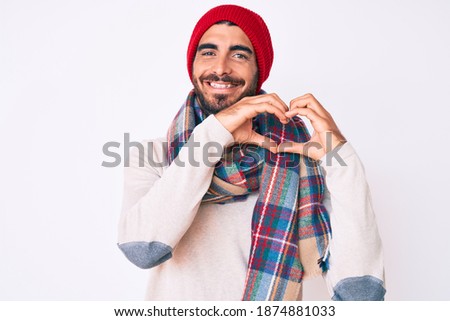 Handsome young man with curly hair and bear wearing a winter sweater, scarf and wool hat smiling in love doing heart symbol shape with hands. romantic concept. 