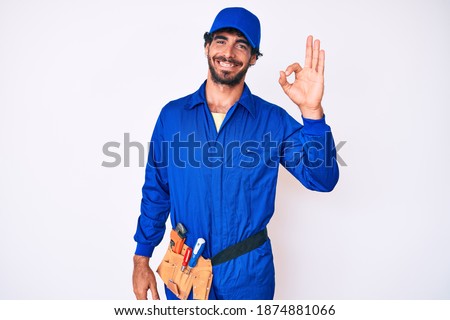 Handsome young man with curly hair and bear weaing handyman uniform smiling positive doing ok sign with hand and fingers. successful expression. 