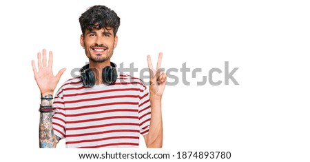 Young hispanic man listening to music using headphones showing and pointing up with fingers number seven while smiling confident and happy. 