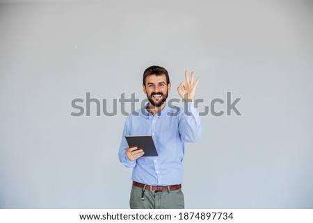 Smiling bearded young CEO standing in front of white background, holding tablet and showing okay sign while looking at camera. The business is going perfectly well.