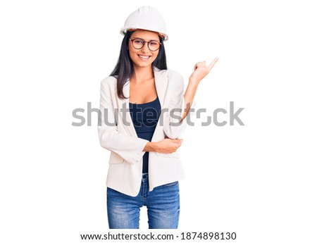 Young beautiful latin girl wearing architect hardhat and glasses with a big smile on face, pointing with hand and finger to the side looking at the camera. 