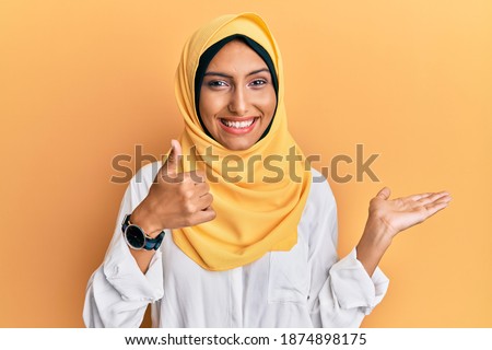 Young brunette arab woman wearing traditional islamic hijab scarf showing palm hand and doing ok gesture with thumbs up, smiling happy and cheerful 