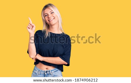 Young beautiful blonde woman wearing casual t-shirt with a big smile on face, pointing with hand and finger to the side looking at the camera. 