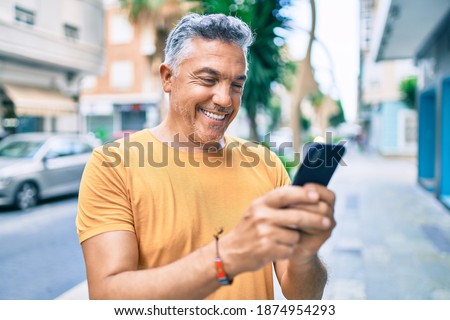 Middle age grey-haired man smiling happy using smartphone walking at street of city. Royalty-Free Stock Photo #1874954293