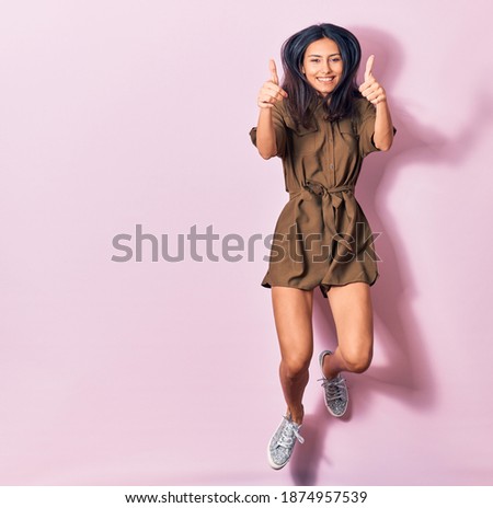 Young beautiful latin woman wearing casual clothes smiling happy. Jumping with smile on face doing ok sign with thumbs up over isolated pink background