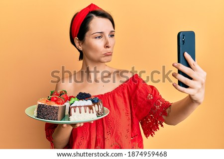 Young brunette woman with short hair holding sweets cakes taking a selfie picture depressed and worry for distress, crying angry and afraid. sad expression. 