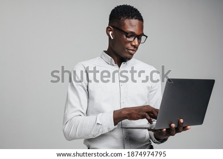 Handsome smiling young african business man in wireless earphones using laptop computer isolated over gray background, wearing white formal shirt