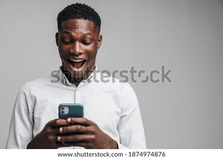 Handsome excited young african business man using mobile phone isolated over gray background, wearing white formal shirt