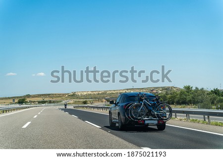 Modern black car with two bicycles attached to the trunk on a straight desert road with blue clear sky and green bushes on a summer day in Spain