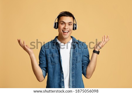 Relax, favorite music, fun and pleasure at spare time. Smiling excited teen student man in casual with modern headphones spreads his hands in sides, isolated on light sandy background, empty space Royalty-Free Stock Photo #1875056098
