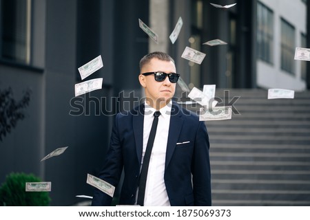 Money rain, falling dollars. Portrait shot of cheerful rich handsome businessman in glasses and suit throws money. Happy good looking man throwing dollars. Royalty-Free Stock Photo #1875069373