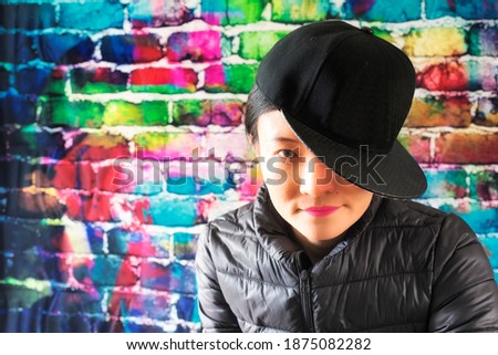 A chinese woman posing against a color brick background wearing a jacket and baseball hat to the side smiling.
