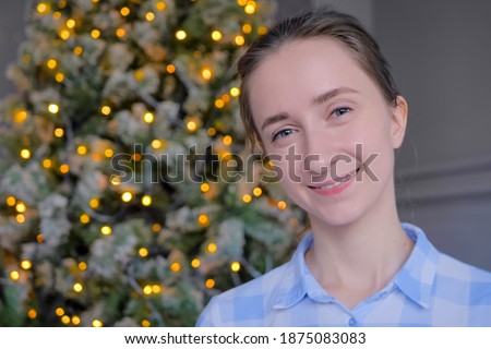 Portrait of young adult positive hipster, student or entrepreneur woman in plaid shirt smiling and looking at camera in room with Christmas tree. Garland light illumination. People and holiday concept