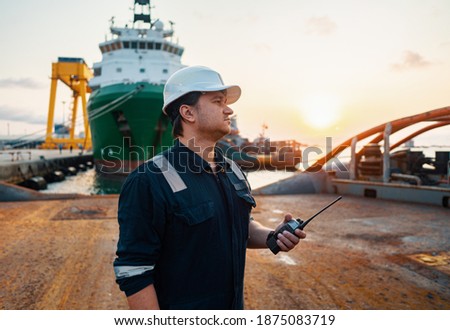 Marine Deck Officer or Chief mate on deck of offshore vessel or ship , wearing PPE personal protective equipment - helmet, coverall. He holds VHF walkie-talkie radio in hands. Royalty-Free Stock Photo #1875083719