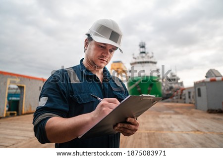 Marine Deck Officer or Chief mate on deck of offshore vessel or ship doing check and filling checklist. Paperwork at sea. Ship is on background Royalty-Free Stock Photo #1875083971