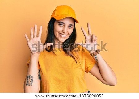 Young hispanic woman wearing delivery uniform and cap showing and pointing up with fingers number eight while smiling confident and happy. 