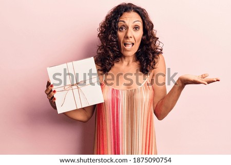 Middle age beautiful woman holding gift celebrating achievement with happy smile and winner expression with raised hand 