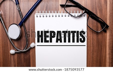 The word HEPATITIS is written on white paper on a wooden background near a stethoscope and black-framed glasses. Medical concept