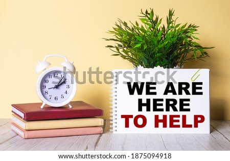 WE ARE HERE TO HELP is written in a notebook next to a green plant and a white alarm clock, which stands on colorful diaries.