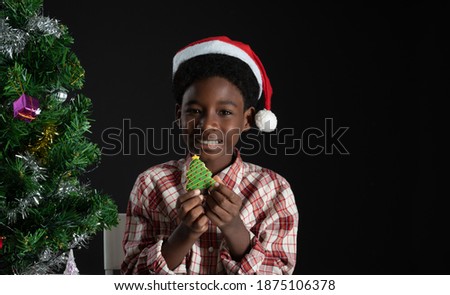 Happy African boy smile and holding Christmas tree shape cookie in hand near little Christmas tree on black background. Christmas New year celebration concept