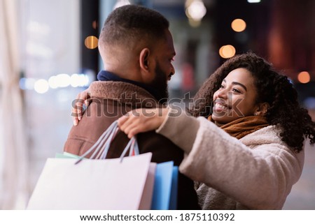 Couple In Love. Cheerful beautiful black lady hugging her boyfriend or husband, holding shopping bags and looking at him. Happy african american family walking near store outdoors in the evening
