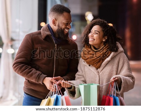 Enjoying Purchases. Portrait of happy smiling black couple holding colorful shopping bags, excited about their new clothes or gift, standing outdoors near mall in the evening, looking at each other