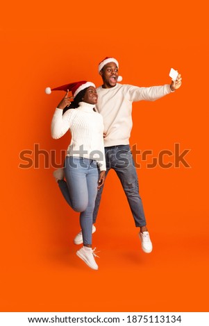 Joyful Black Couple Taking Selfie On Smartphone, Having Fun On Christmas Holidays, Jumping And Posing On Orange Studio Background, Enjoying Celebrating Xmas Together, Full Length Shot, Copy Space