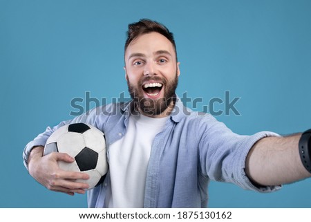 Happy soccer fan with ball taking selfie on mobile device over blue studio background. Positive football rooter making photo of himself after game, cheering for his favorite team