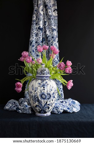 Lush natural light old Dutch Delft blue lidded vase with pink tulips in the background on navy blue linen against a black background.
