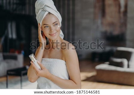 Pleased lovely young European woman applies body lotion wrapped in bath towel looks away with thoughtful expression stands indoor against blurred background in living room. Skin care concept