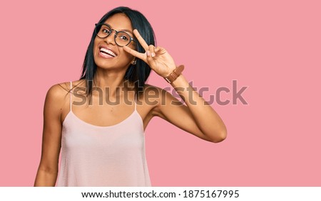 Young african american woman wearing casual clothes and glasses doing peace symbol with fingers over face, smiling cheerful showing victory 