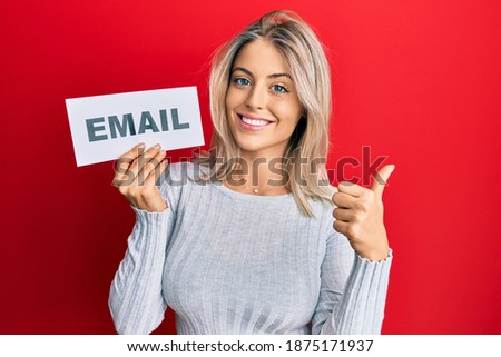 Beautiful blonde woman holding paper with email address smiling happy and positive, thumb up doing excellent and approval sign 