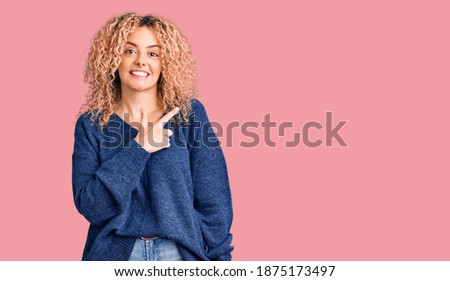 Young blonde woman with curly hair wearing casual winter sweater cheerful with a smile on face pointing with hand and finger up to the side with happy and natural expression 