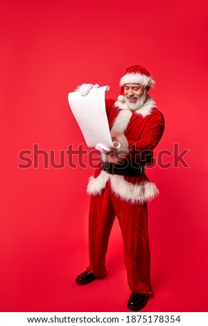 Santa Claus wearing costume holding parchment roll reading letter wish list, elderly male is preparing for Christmas holiday standing isolated on red background