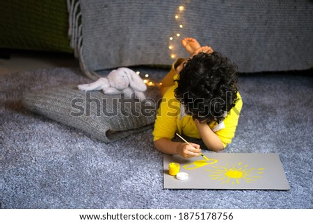Little curly boy in yellow clothes painting sun in gray interior. 