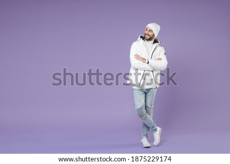 Full length of smiling man in warm white padded windbreaker jacket hat holding hands crossed looking aside isolated on purple background studio portrait. People lifestyle cold winter season concept