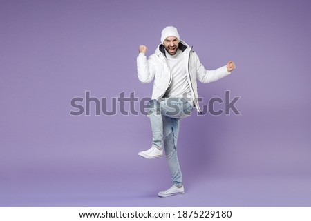 Full length of overjoyed man in white windbreaker jacket hat doing winner gesture clenching fist celebrating isolated on purple background studio portrait. People lifestyle cold winter season concept