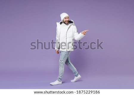 Full length side view of excited man in warm white windbreaker jacket hat pointing index finger aside isolated on purple color background studio portrait. People lifestyle cold winter season concept